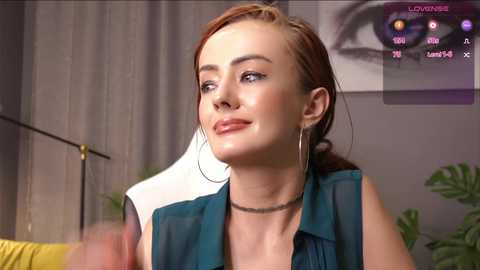 Media: Video of a young woman with fair skin, light brown hair, wearing a teal blouse, silver hoop earrings, and a choker necklace, indoors near a plant.