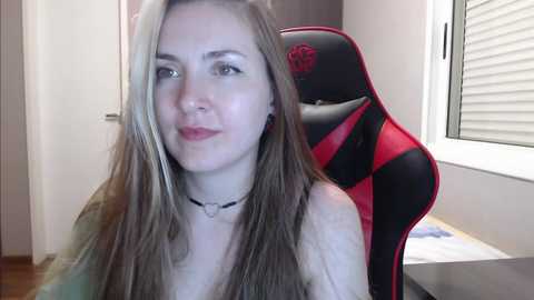 Media: Video of a fair-skinned, blonde-haired woman with a light makeup look, wearing a black choker, sitting in a red and black gaming chair. Background shows a window with white blinds and a desk with scattered papers.