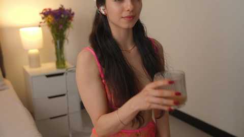 Media: Video of a slender, fair-skinned woman with long black hair in pigtails, wearing a pink tank top, holding a glass of wine, in a modern bedroom with white furniture and a vase of purple flowers.