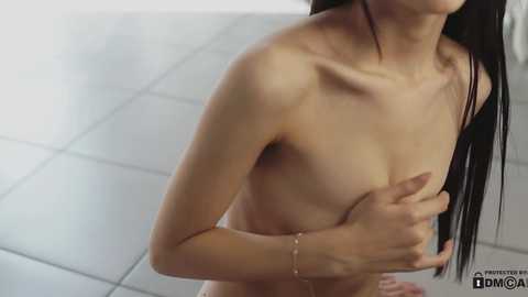 Media: A video of a nude, slim Asian woman with long black hair, covering her small breasts with one hand, standing on a tiled floor. She wears a delicate bracelet on her left wrist.