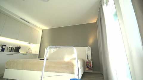 Media: Video of a minimalist, modern bedroom with beige walls, white furniture, and a transparent chair in the foreground; a bed with beige bedding, a white dresser, and a small bookshelf with books and a lamp on the left.