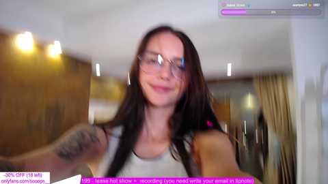 Media: Video of a young woman with long brown hair, glasses, and a white tank top, smiling and waving, indoors with a blurred background.