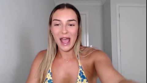 Media: A video of a young Caucasian woman with light skin and long, straight blonde hair. She's wearing a yellow and blue patterned bikini top, sticking out her tongue and smiling. The background is a plain white wall and door.