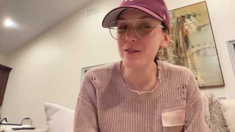 Media: Video of a fair-skinned woman with glasses and a maroon cap, wearing a beige knit sweater with a small pocket, seated in a modern bedroom with a white bed and framed artwork on the beige walls.