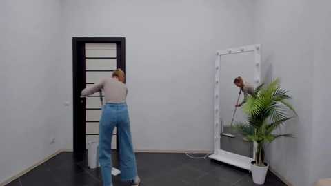 Media: Video of a blonde woman in a beige sweater and blue jeans, standing with her back to a tall mirror, adjusting a vertical door with horizontal blinds in a minimalist, white-walled room with dark flooring and a green potted plant.