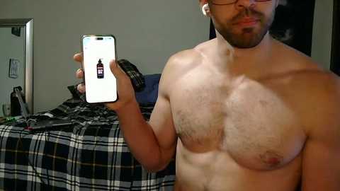 Media: A shirtless man with a beard, holding a smartphone displaying a bottle of whiskey, stands in a dimly-lit bedroom with a plaid-patterned bedspread and a partially visible mirror.