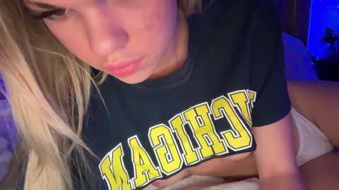 Media: Video of a blonde woman with fair skin, wearing a black \"Wabash\" t-shirt, lying on a bed, visible cleavage, dimly lit room.