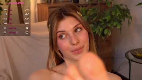 Media: A video of a young woman with light skin and shoulder-length brown hair, wearing a black bra, in a cozy, dimly lit room with a potted plant and wooden furniture.