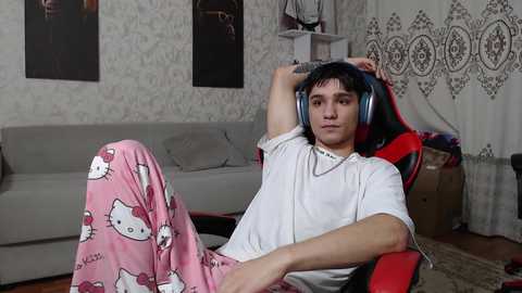 Media: Video of a young man in white t-shirt and pink Hello Kitty pajama pants, wearing large headphones, relaxing in a red gaming chair in a cozy living room.