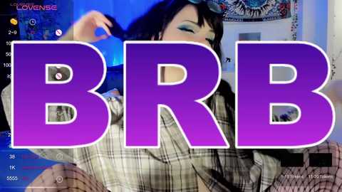 Media: Video of a woman with dark hair, wearing sunglasses, holding a black hair tie. She's in a plaid shirt, with the word \"BRB\" in bold purple letters overlaying her. Background shows a blue room with a tapestry.