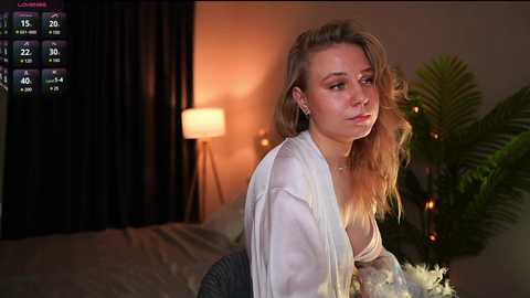 Media: Video of a young Caucasian woman with blonde hair, wearing a white blouse and grey skirt, leaning forward in a dimly-lit bedroom with a modern lamp and large potted plant.