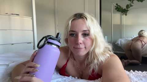 Media: Video of a young, blonde woman with fair skin lying on a bed, wearing a red bra, holding a lavender water bottle. Background shows a white dresser and a partially visible mirror reflecting a green plant.