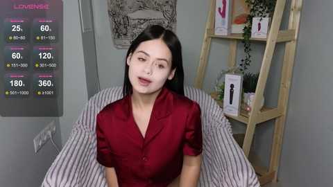 Media: Video of a young woman with long black hair and fair skin, wearing a red button-up shirt, seated on a striped bed. Background includes a wooden ladder bookshelf with various products, a black and white tapestry, and a digital display with health metrics.