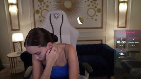 Media: Video of a young woman with fair skin and dark hair, wearing a blue tank top, sitting in a white gaming chair in a dimly lit, luxurious room with a plush dark blue couch and decorative wall art.