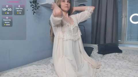 Media: Video of a young, slender woman with fair skin and long blonde hair in a white, frilly nightgown, posing in a modern bedroom with light walls, a bed, and a window.