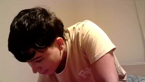 Media: Video of a young woman with short, dark hair and pale skin, wearing a beige t-shirt, bending forward in a simple, beige-walled room.