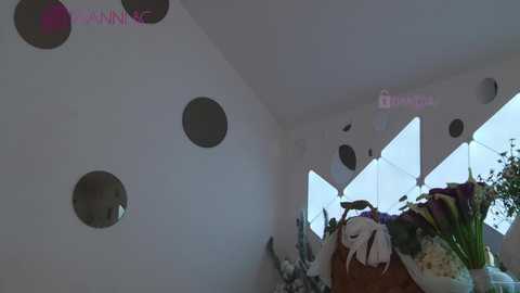 Media: Video of a modern room with a white wall featuring circular cutouts and a large window with diamond-shaped panes. A vase of white flowers sits on a wooden table in the foreground.