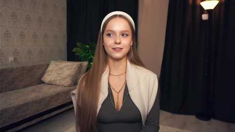 Media: Video of a young, fair-skinned woman with long brown hair, wearing a white headband, beige blazer, and black top, sitting in a modern living room with beige couch, green plant, and dark curtains.