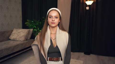 Media: Video of a young Caucasian woman with long, straight brown hair, wearing a beige headscarf, black top, and beige cardigan, standing in a modern living room with gray couch, green plant, and dark curtains.