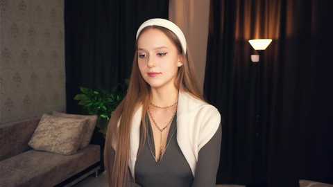 Media: Video of a young Caucasian woman with long, straight, light brown hair, wearing a white headband, dark dress, and gold necklace, standing in a dimly lit, modern living room with dark curtains and a soft-lit wall lamp.