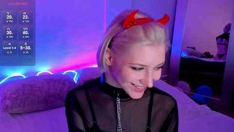 Media: Video of a young, fair-skinned woman with blonde hair, wearing a black sheer top and red devil horns, smiling. Neon lights and a TV screen with a calendar in the background.