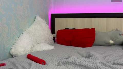 Media: A video of a neatly made bed with a plush white pillow, red pillow, and gray blanket. A red vibrator lies on the bed, against a light blue, patterned wallpaper and a wooden headboard.