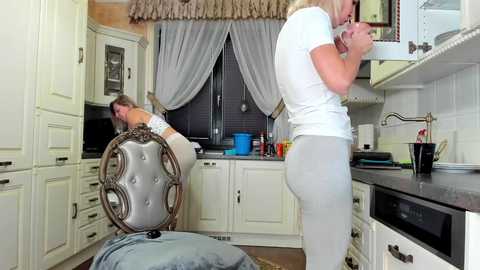 Media: Video of a blonde woman in a white top and gray pants standing in a modern, white kitchen with gray countertops, a sink, and cabinets. A second woman in a white top and jeans sits on a chair, peering under it.