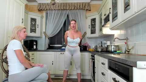 Media: Video of a modern kitchen with white cabinets and beige walls. Two blonde women, one in a white T-shirt, the other in a blue top and leggings, stand facing each other.