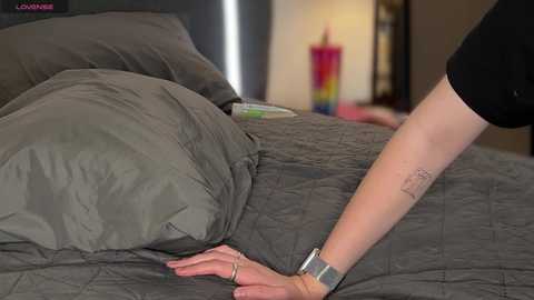 Media: Video of a person wearing a black shirt, lying on a gray quilted bedspread, with a tattooed arm reaching out. Background includes a colorful candle and a blurred person.