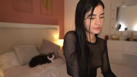 Media: Video of a young woman with long black hair, wearing a black mesh top, standing in a softly lit bedroom with a white bed, white dresser, and a black-and-white cat.