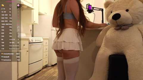 Media: Video of a young woman in a white skirt and thigh-high stockings, facing a teddy bear in a cozy kitchen with a stove, wooden floor, and a smartphone recording.