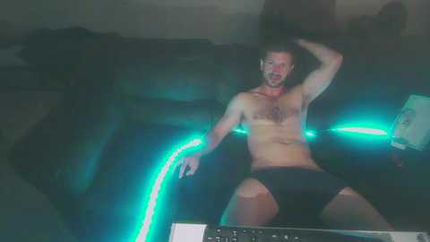 Media: A video shows a shirtless, muscular man with fair skin and short hair, wearing only black briefs, sitting on a dark couch. He has an open-mouthed expression, with glowing neon lights in the background.