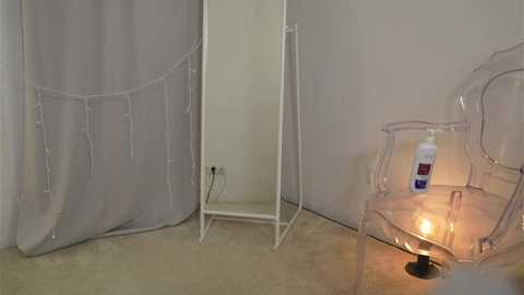 Media: Video of a minimalist, white-walled room with a clear plastic chair, white curtain, and a lit candle on a small table.