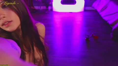 Media: Video of a nude woman with long brown hair, small breasts, and light skin, taking a selfie in a dimly lit room with purple lighting and a glowing white cube.