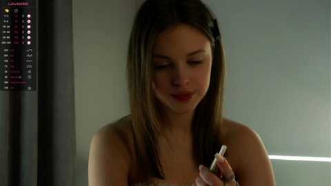 Media: Video of a young woman with long brown hair, fair skin, and a slender build, holding a white pen, in a dimly lit room with a thermometer on the wall.