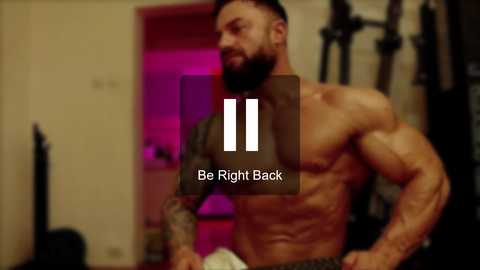 Media: Video of a shirtless, muscular man with a beard and tattoos, flexing in a gym with dim lighting, overlaid with \"Be Right Back\" text and a vertical line icon.