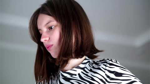 Media: Video of a young woman with straight, shoulder-length brown hair and a zebra-patterned shirt, gazing thoughtfully to the side. The background is a blurred, neutral tone.