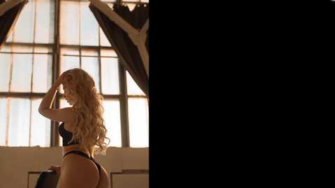 Media: Video of a curvaceous blonde woman with wavy hair, wearing a black bra and thong, standing in a dimly lit room with large arched windows and drapes.