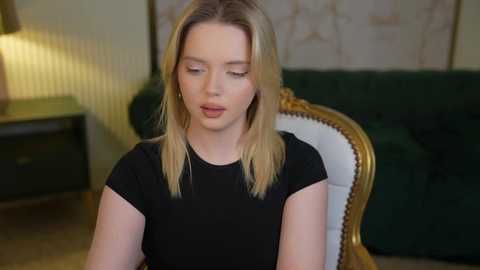 Media: Video of a blonde woman with fair skin, wearing a black top, sitting on an ornate chair with a green velvet background.