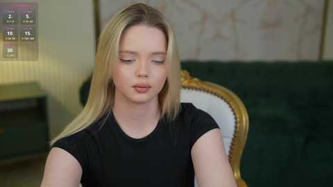 Media: Video of a young Caucasian woman with long blonde hair, wearing a black top, sitting on a green velvet couch with a golden frame. Background shows a beige wall with a subtle floral pattern.