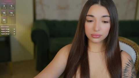 Media: Video of a young woman with long, straight brown hair and a medium skin tone, seated indoors. She has a slender build, and a tattoo on her left shoulder. Background includes a green sofa and a blurred wall.