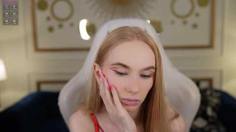 Media: Video of a young blonde woman with long hair, wearing a white veil and red dress, applying makeup with pink nail polish, against a blurred, elegant interior background.