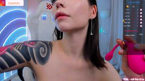 Media: Video of a light-skinned woman with dark hair, piercings, and a colorful tattoo on her right shoulder, holding a pink vibrator. Background includes a blue and white abstract light display and a social media logo.