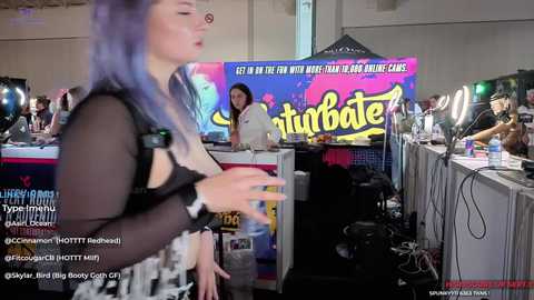 Media: Video of a woman with purple hair, wearing a sheer black top, speaking at a convention booth. Background features vibrant \"Turbote\" branding and attendees.