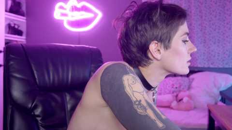 Media: Video of a topless woman with short dark hair, tattoos, and a black choker, sitting on a black leather chair, in a neon-lit room with a pink lipstick sign and floral wallpaper.