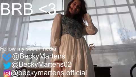 Media: Video of a young woman with dark hair in a white and blue floral dress, standing in front of a window. Text overlay with social media handles.