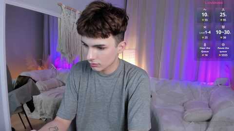 Media: Video of a young Caucasian boy with short brown hair, wearing a gray t-shirt, sitting in a dimly lit room with a cream sofa, purple lights, and a macrame wall hanging.