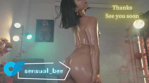 Media: Video of a nude, dark-skinned woman with shoulder-length black hair, backside prominently displayed, standing in a dimly lit room with pink and blue lights, surrounded by floral decor.