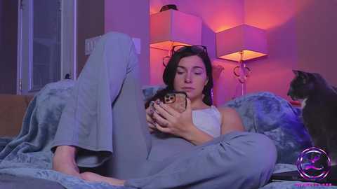 Media: Video of a woman with long dark hair, wearing a white tank top and gray blanket, reading on a bed under purple lights, with a black cat beside her.