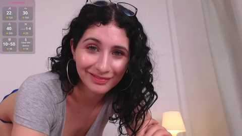 Media: Video of a smiling, curly-haired woman with medium skin tone, wearing glasses, a grey T-shirt, and large hoop earrings, in a modern, white room with a digital clock and lamp.